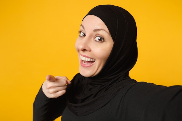 Cerca de joven musulmana árabe en ropa negra hijab haciendo selfie disparo en teléfono móvil aislado en retrato de pared amarilla. Concepto de estilo de vida religioso de la gente.