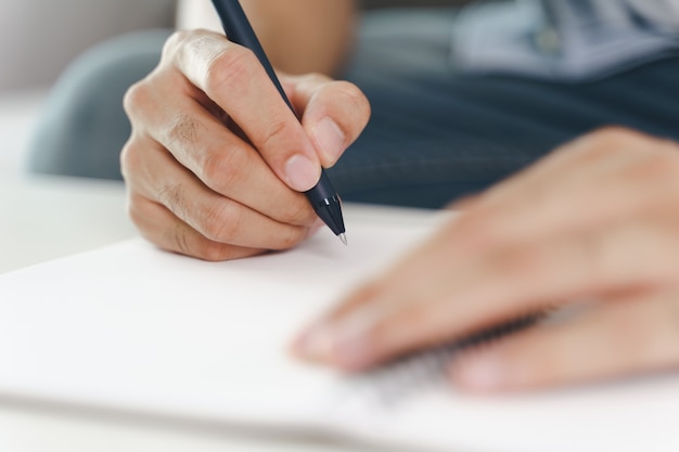 Cerca de joven en paño casual manos escribiendo en el bloc de notas, cuaderno con bolígrafo sobre la mesa.