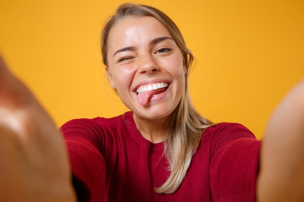 Foto cerca de una joven rubia divertida con ropa informal posando aislada en un fondo amarillo anaranjado. concepto de estilo de vida de las personas. simulacros de espacio de copia. haciendo selfie en el teléfono móvil mostrando la lengua.