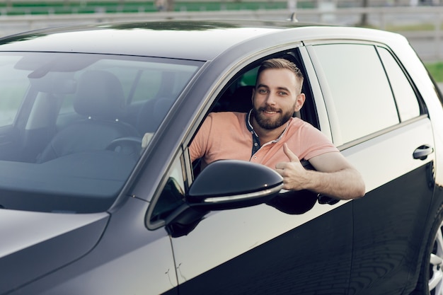 Cerca de joven sonriendo y mostrando los pulgares para arriba en su nuevo coche. Comprando un carro.