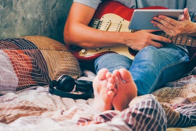 Cerca de joven usando tableta y tocando la guitarra eléctrica en su cama