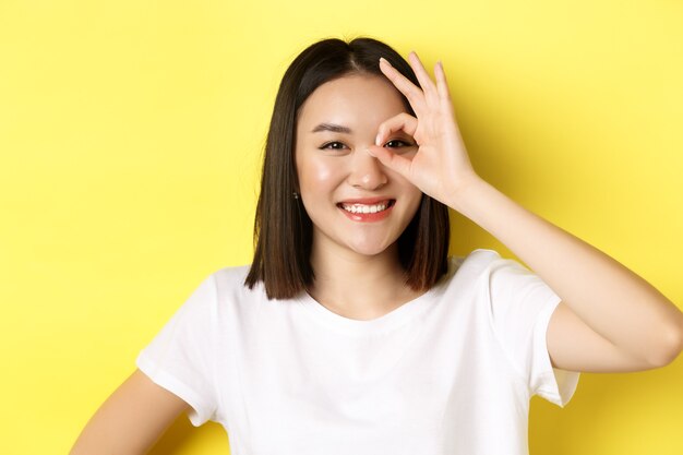 Cerca de una linda chica asiática que se siente feliz, mostrando el signo de OK en el ojo y sonriendo, de pie sobre el amarillo.