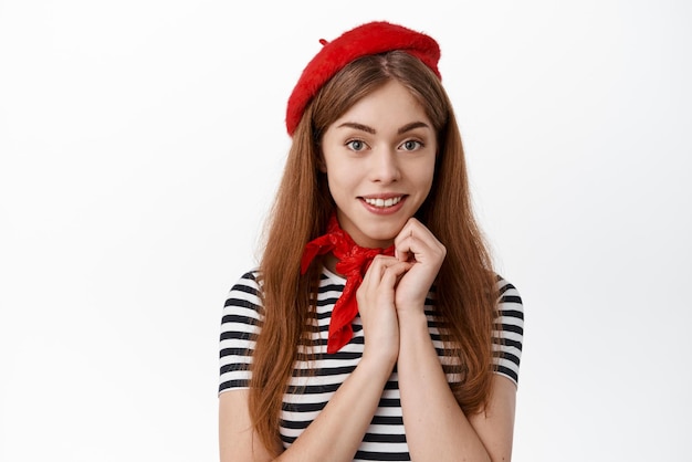 Foto cerca de una linda estudiante con ropa de sombrero francés sonriendo y mirando linda a la cámara de pie contra el fondo blanco
