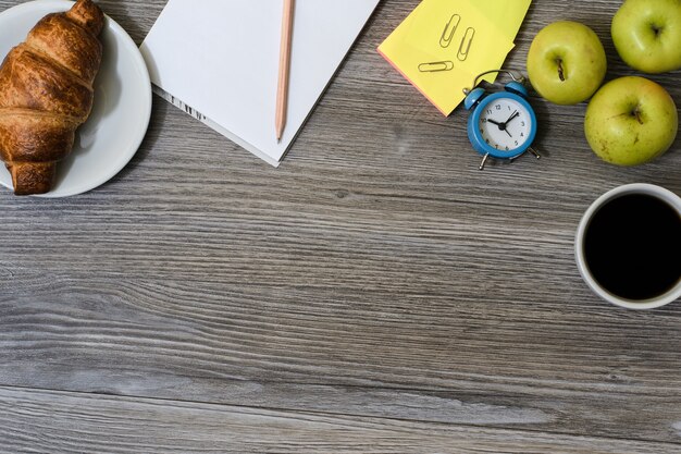 Foto cerca del lugar de trabajo: taza de café, manzanas, plato con croissant, bloc de notas y lápiz, reloj despertador y una mesa