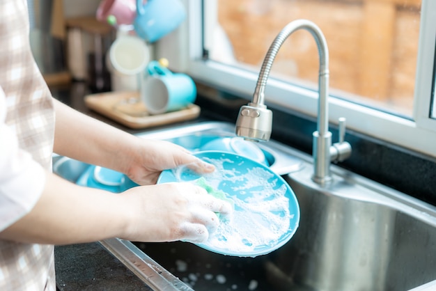 Cerca de la mano de la atractiva joven asiática está lavando platos en el fregadero de la cocina mientras hace la limpieza en casa durante su estancia en casa utilizando el tiempo libre sobre su rutina diaria de limpieza.