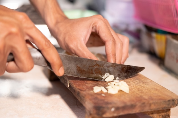 Cerca de la mano del cocinero mientras corta el ajo con un cuchillo y una tabla de cortar de madera