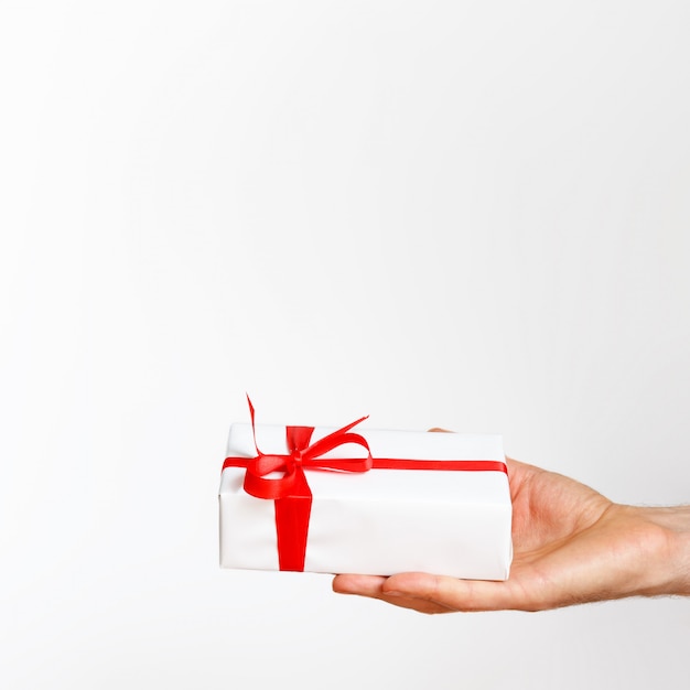 Cerca de la mano del hombre sosteniendo una caja de regalo envuelta en blanco con cinta roja, aislado sobre fondo blanco, vista lateral.