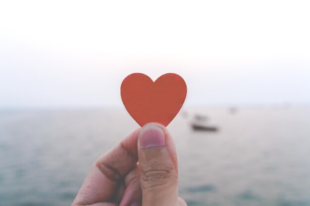 Cerca de la mano del hombre sosteniendo un corazón de papel con el fondo del mar concepto de amor feliz día de San Valentín