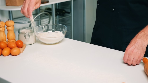 Cerca de la mano del hombre tamizando la harina de trigo en la mesa del comedor. Panadero senior jubilado con delantal, uniforme de cocina rociando, tamizando, extendiendo ingredientes rew para hornear pan y pizza casera.