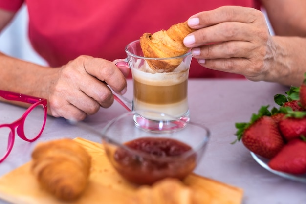 Cerca de la mano humana de la mujer madura disfrutando del desayuno en casa con croissant capuchino fresco