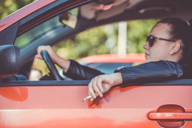 Cerca de la mano de la mujer fumando cigarrillo dentro del automóvil mientras conduce un vehículo, conducción amenazadora y peligrosa