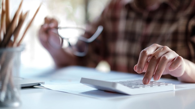 Cerca de la mano de la mujer o del contador que sostiene la pluma trabajando en la calculadora para calcular datos comerciales, documentos contables y computadora portátil en la oficina