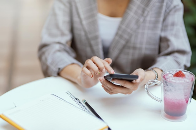 Foto cerca de una mano de mujer sosteniendo un teléfono móvil un vaso de néctar colocado sobre la mesa en el café