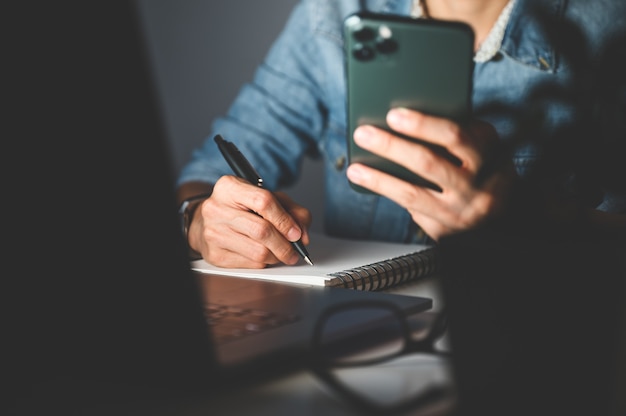 Cerca de la mano de la mujer con teléfono celular o móvil para comprar en línea o pagar y escribir algo en el cuaderno. Concepto de compras en línea.