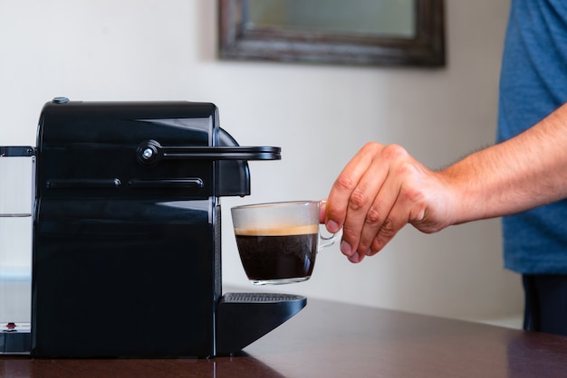 Cerca de la mano recogiendo una taza de café espresso de la máquina de café de cápsulas en casa.