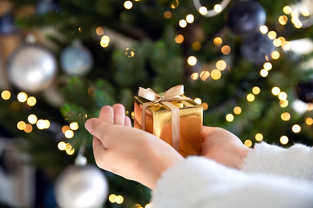 Cerca de mano sosteniendo una caja de regalo dorada con árbol de Navidad y luces