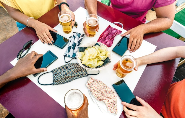 Cerca de las manos de amigos cerca de mascarillas en la mesa con teléfonos inteligentes móviles y cervezas