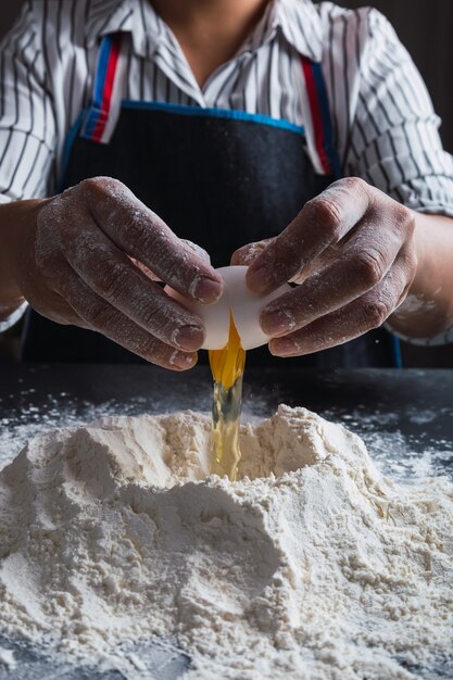 Cerca de las manos del cocinero rompiendo el huevo en la harina.