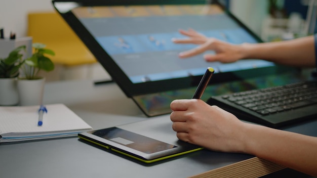 Cerca de las manos del especialista en medios usando tableta gráfica con lápiz y pantalla táctil en el monitor para software de retoque fotográfico en el estudio. Editor de imágenes trabajando con equipos de retoque.
