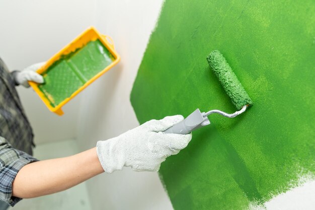 Cerca de las manos en guantes de trabajo pintando una pared verde con un rodillo de tamaño medio