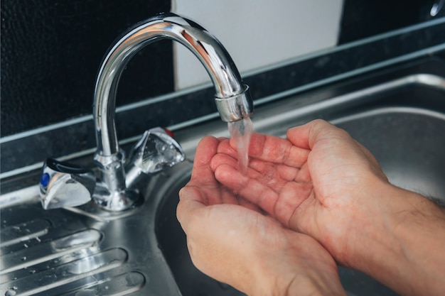 Cerca de las manos masculinas que se lavan en un fregadero de metal. Un juego de agua en la palma de tu mano para lavarte la cara. Procedimiento higiénico matutino. Prevención de virus y enfermedades. Eliminación de microbios.