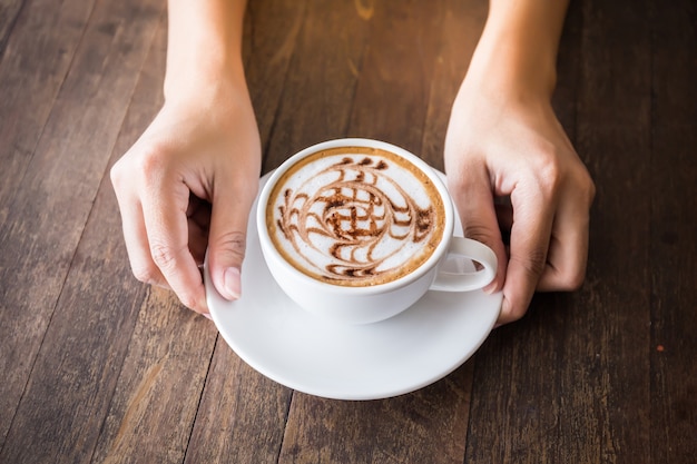 Cerca de las manos de mujer sosteniendo la taza de café blanco con arte hermoso patrón en la mesa de madera