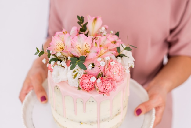 Cerca de las manos de las mujeres sosteniendo un pastel decorado con flores sobre un fondo blanco.