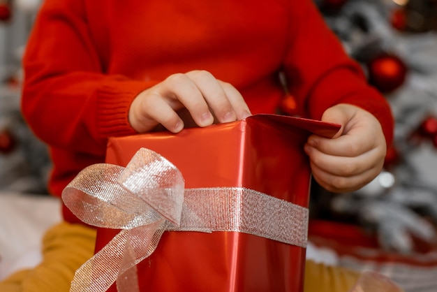 Foto cerca de las manos del niño pequeño abriendo regalo de navidad con lazo blanco sobre fondo de árbol de navidad