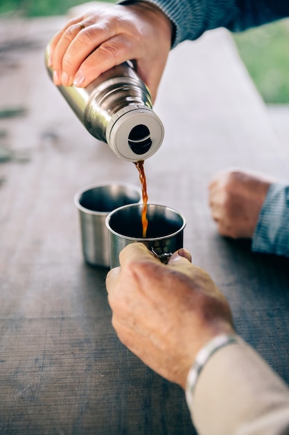 Cerca de las manos de la pareja senior vertiendo café en una taza de metal del termo sobre una mesa de madera