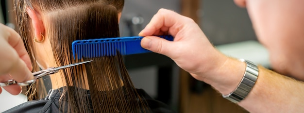 Cerca de las manos del peluquero masculino cortando el cabello largo de la mujer joven con tijeras y peine en el salón