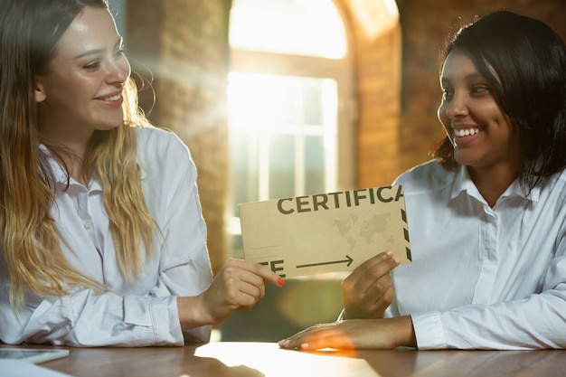 Cerca de las manos del ser humano afroamericano y caucásico con certificado o tarjeta de invitación. Mujeres felices, sonrientes y lindas. Negocios, trabajo en equipo, estudio, educación, finanzas, concepto festivo.