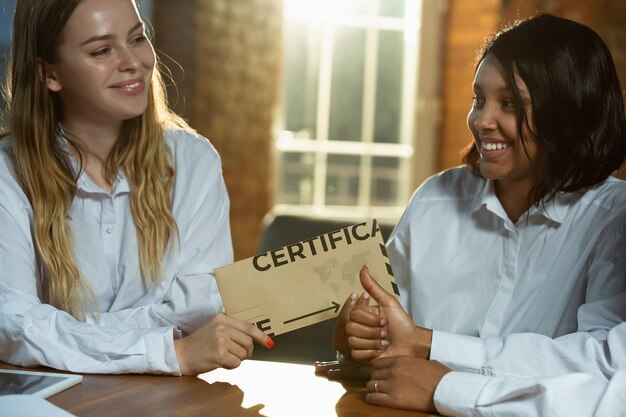 Cerca de las manos del ser humano afroamericano y caucásico con certificado o tarjeta de invitación. Mujeres felices, sonrientes y lindas. Negocios, trabajo en equipo, estudio, educación, finanzas, concepto festivo.