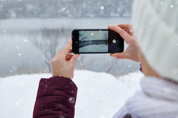 Cerca de manos sosteniendo el teléfono inteligente y haciendo un rodaje de la naturaleza mientras cae nieve