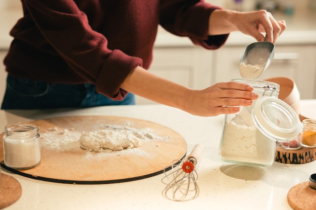 Cerca de la mesa con la masa en la tabla de cortar y la mujer tomando cuidadosamente la harina de la jarra