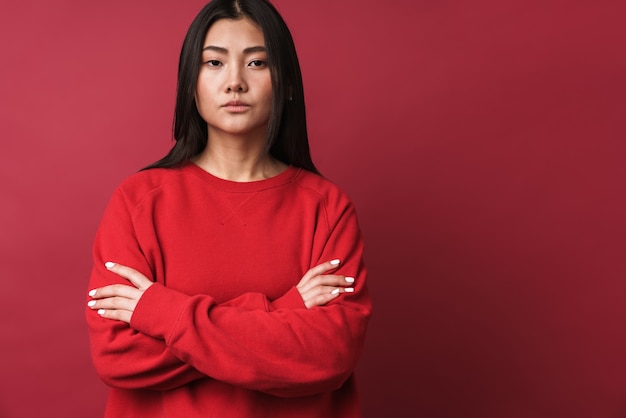 Cerca de una mujer asiática bastante joven con ropa casual que se encuentran aisladas sobre la pared roja, con los brazos cruzados