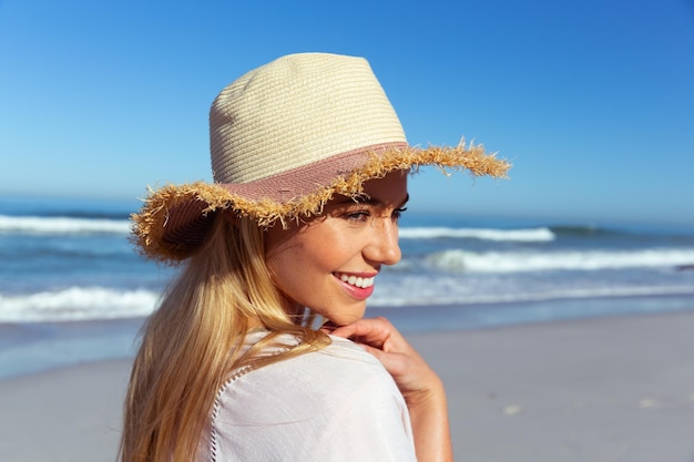 Cerca de una mujer caucásica disfrutando del tiempo en la playa en un día soleado, con un sombrero, sonriendo, con el mar de fondo