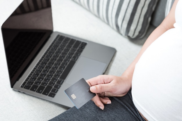 Cerca de una mujer embarazada joven feliz usando una computadora portátil para hacer compras en línea con tarjeta de crédito