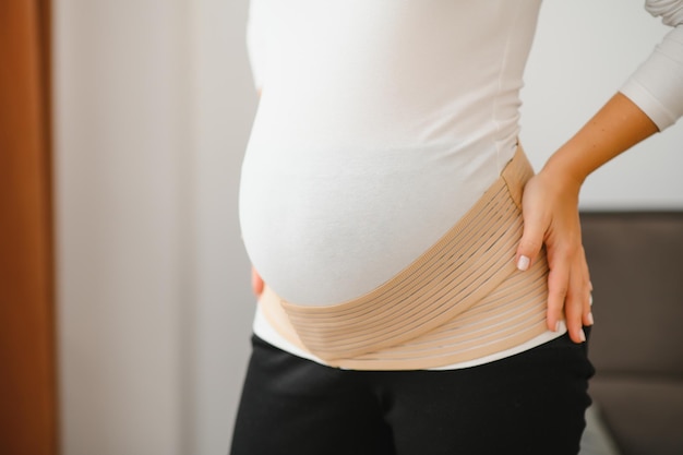 Cerca de la mujer embarazada poniéndose un vendaje en un fondo gris con espacio de copia. Concepto de cinturón de soporte abdominal ortopédico.