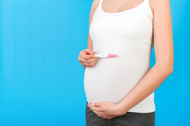 Cerca de la mujer embarazada en ropa de casa con prueba de embarazo positiva en fondo azul. Concepto de maternidad. Copie el espacio.