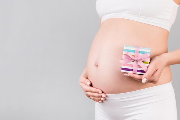 Cerca de la mujer embarazada en ropa interior blanca sosteniendo una caja de regalo en fondo gris. Esperando parto. Copie el espacio.
