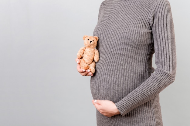 Cerca de la mujer embarazada en vestido gris sosteniendo un oso de peluche contra su vientre en fondo gris. La madre joven está esperando un bebé. Copie el espacio.