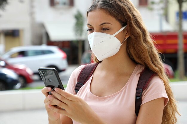 Cerca de la mujer estudiante con mensajes de máscara protectora con teléfono inteligente en las calles de la ciudad