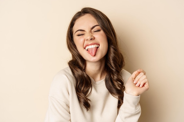 Cerca de una mujer feliz y despreocupada mostrando la lengua y divirtiéndose posando en suéter sobre fondo beige