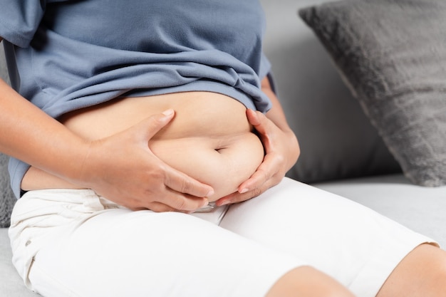 Cerca de la mujer gordita sosteniendo su grasa del vientre en el sofá. Estilo de vida de dieta para reducir el vientre y dar forma a un concepto de músculo abdominal saludable.