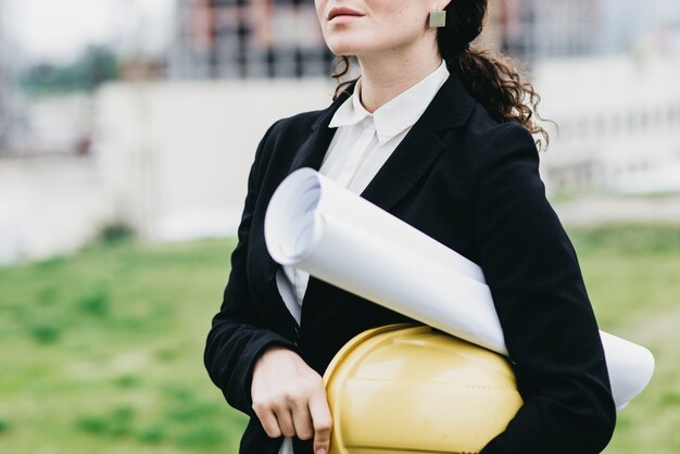 Cerca de mujer ingeniero con casco amarillo en la mano contra el fondo de la construcción de la ciudad