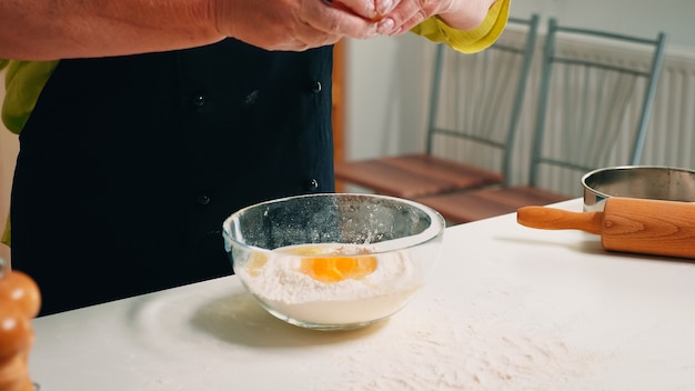 Foto cerca de la mujer de las manos agregando huevos en harina para hornear. anciano chef de repostería agrietando el huevo en un tazón de vidrio para la receta de la torta en la cocina, mezclando a mano, amasando los ingredientes preparando la torta casera