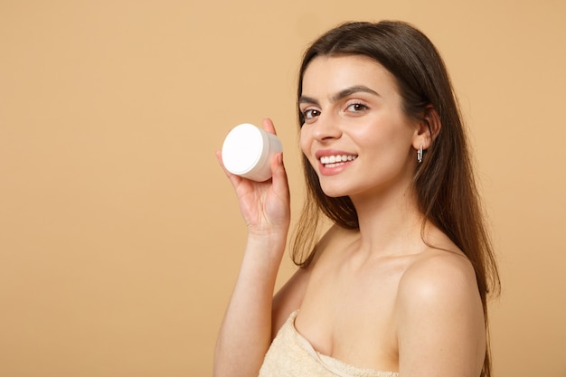 Cerca de mujer medio desnuda con piel perfecta maquillaje desnudo aplicando crema facial aislada en la pared de color beige pastel