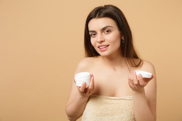 Foto cerca de mujer medio desnuda con piel perfecta maquillaje desnudo aplicando crema facial aislada en la pared de color beige pastel