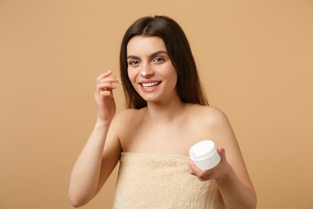 Foto cerca de mujer medio desnuda con piel perfecta maquillaje desnudo aplicando crema facial aislada en la pared de color beige pastel