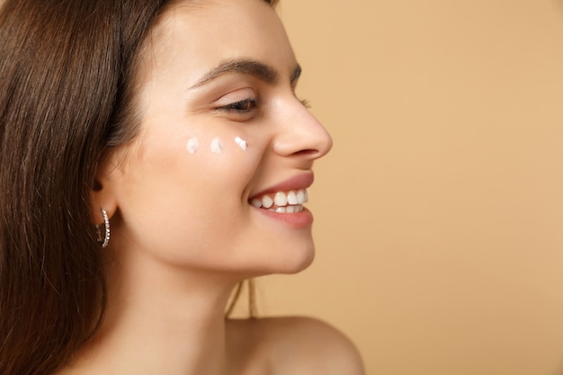 Cerca de mujer medio desnuda con piel perfecta maquillaje desnudo aplicando crema facial aislada en la pared de color beige pastel